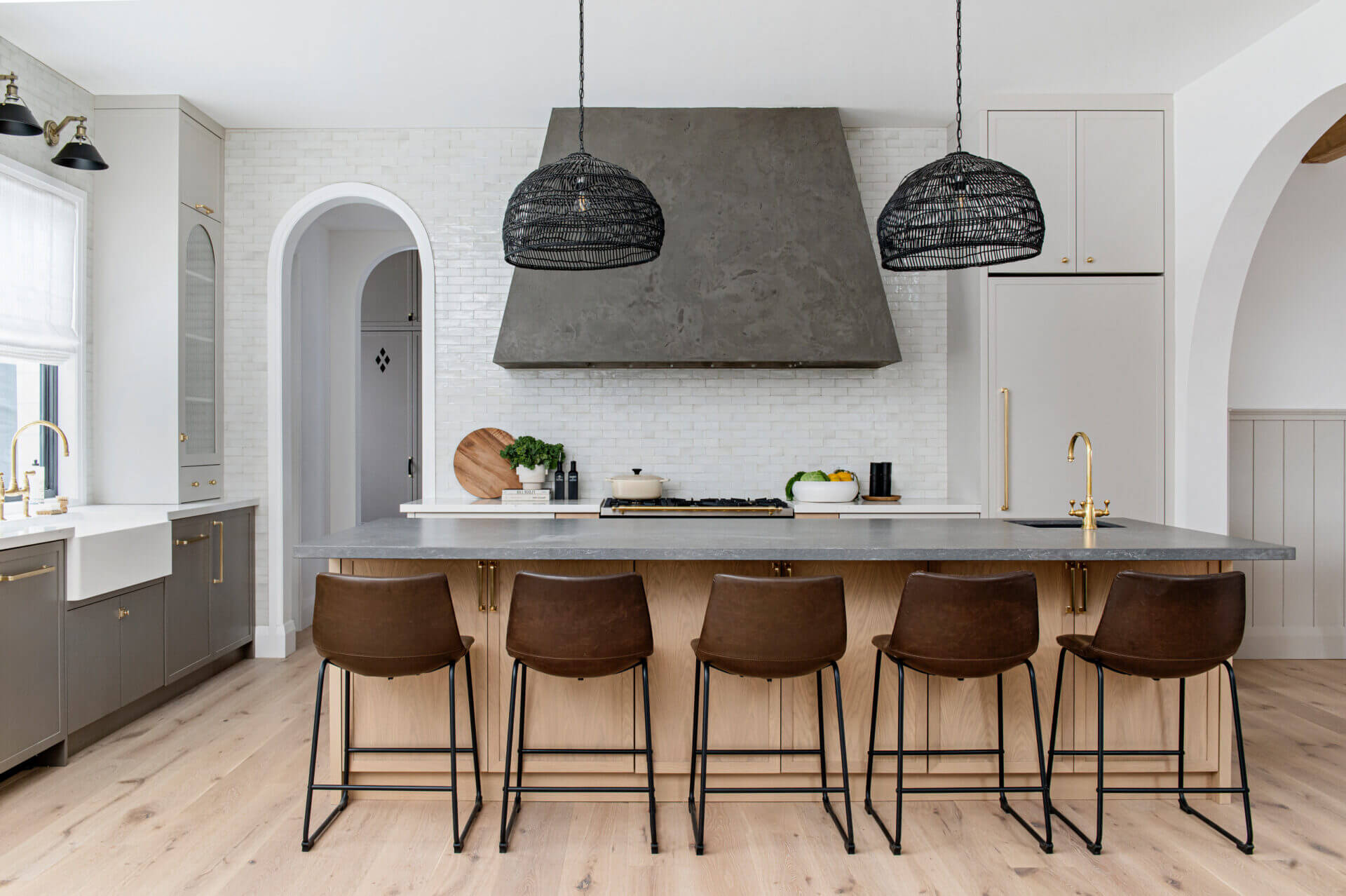 A kitchen with a concrete-look hood, black rattan pendants, brown leather stools and an arched doorway
