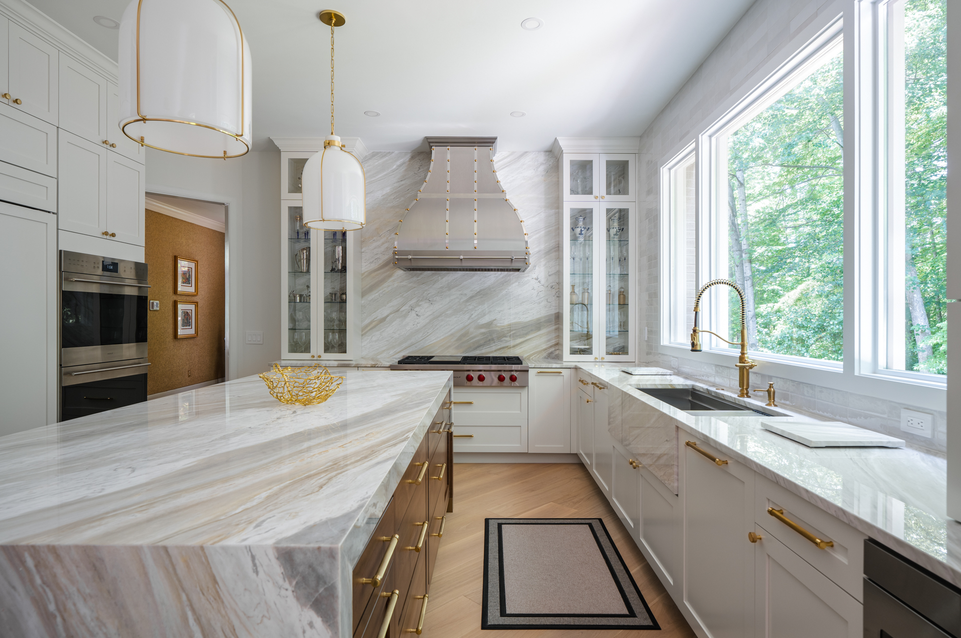 A kitchen with a veined stone island and hood, brass pendants and white cabinetry