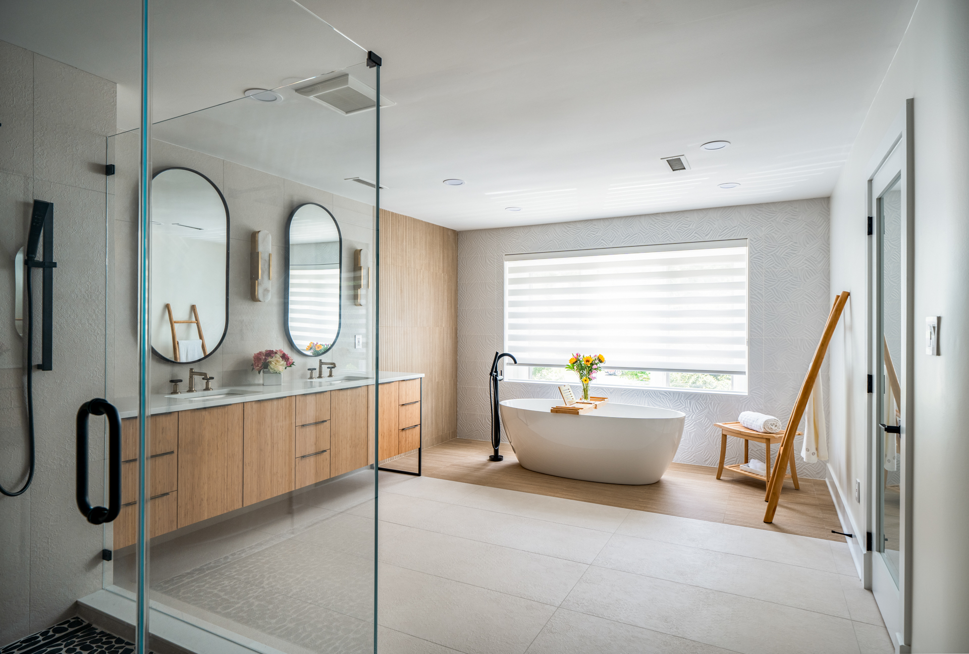 A bathroom seen through glass, with oval mirrors, a soaking tub and an oak vanity